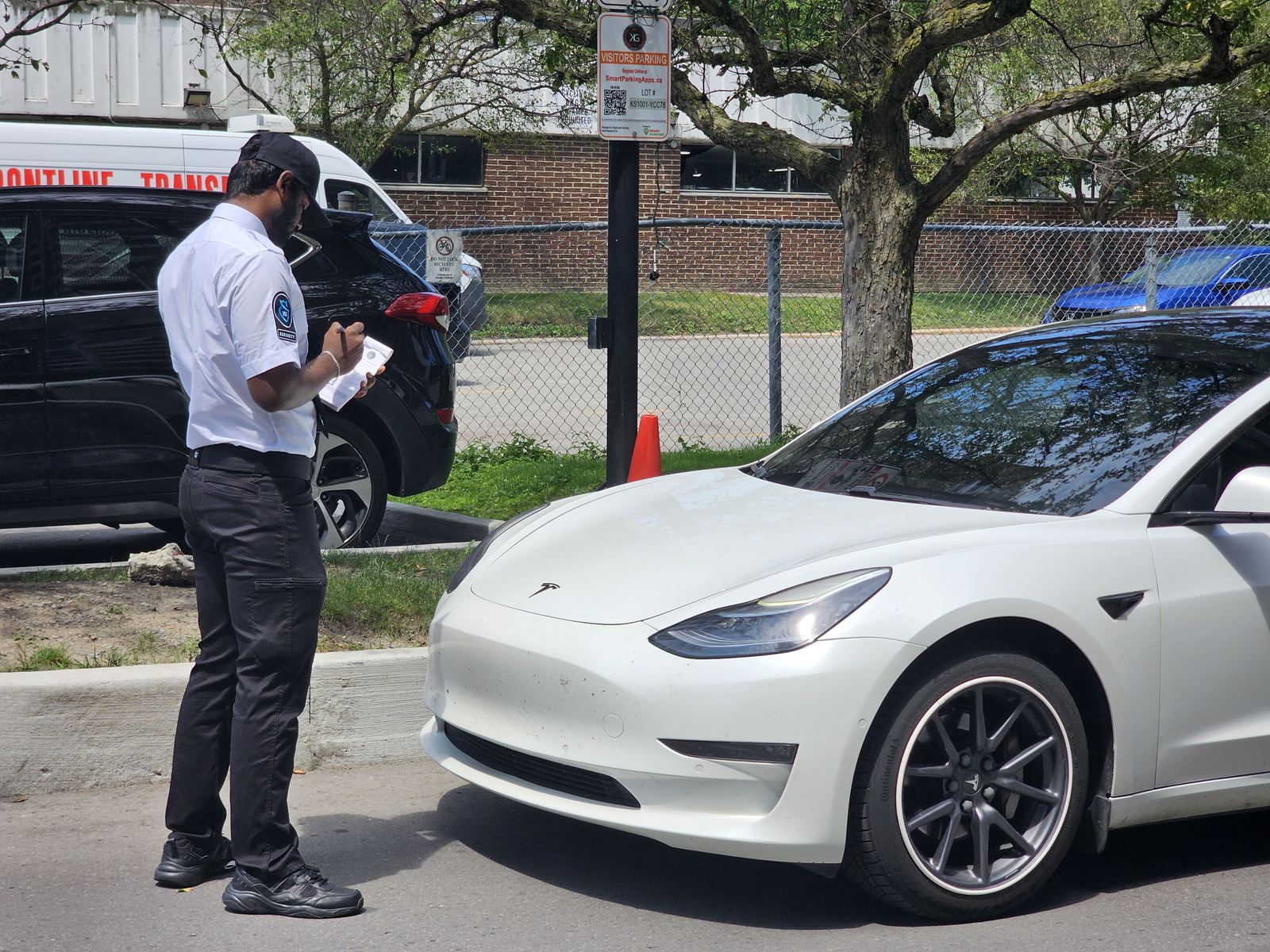 A KingsGuard guard writing a parking notice beside a white car in a visitor parking lot marked with a permit-parking sign