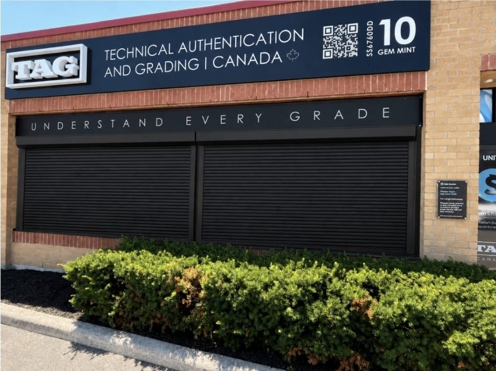Black roll-down security shutters closed across the storefront of TAG Canada, a trading card grading company, below its illuminated sign.