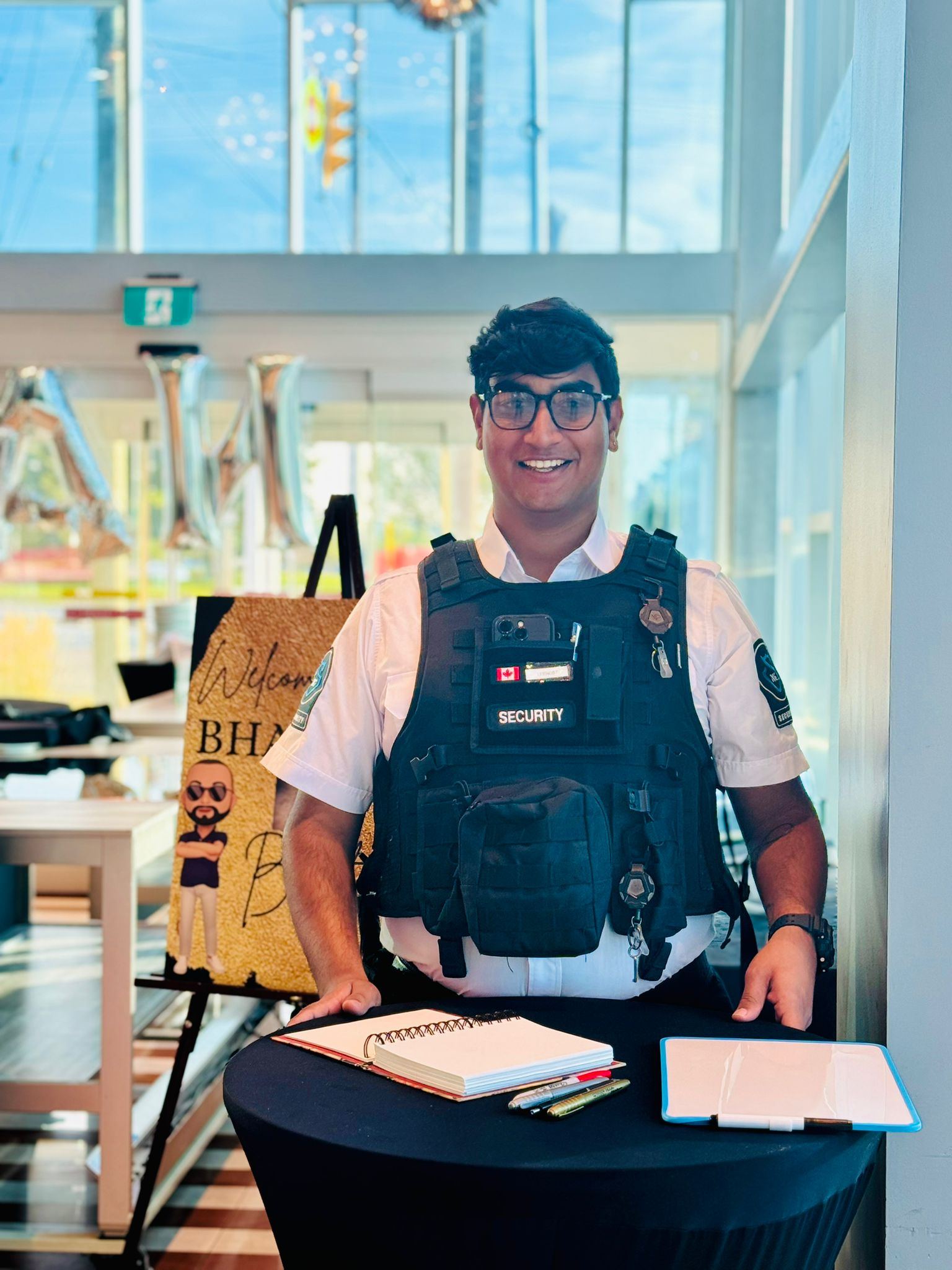 A smiling KingsGuard guard in a white uniform shirt and protective vest standing at a private-event welcome table